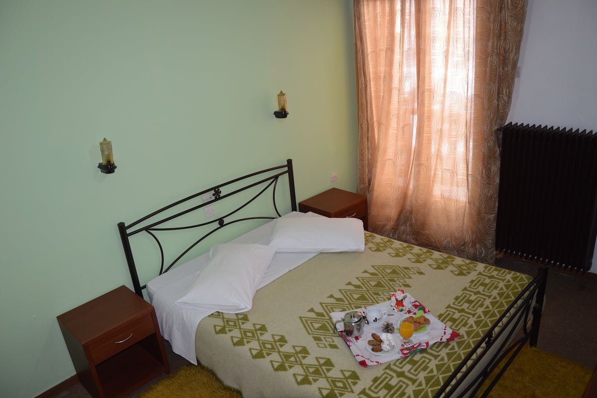Una cama con colcha estampada, una mesita de noche, una lámpara de pared y una ventana con cortinas.