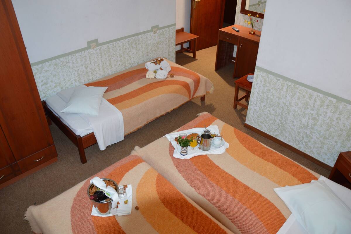 Habitación de hotel con dos camas individuales, un escritorio de madera, un baño con espejo y una pequeña mesa con alimentos para el desayuno.