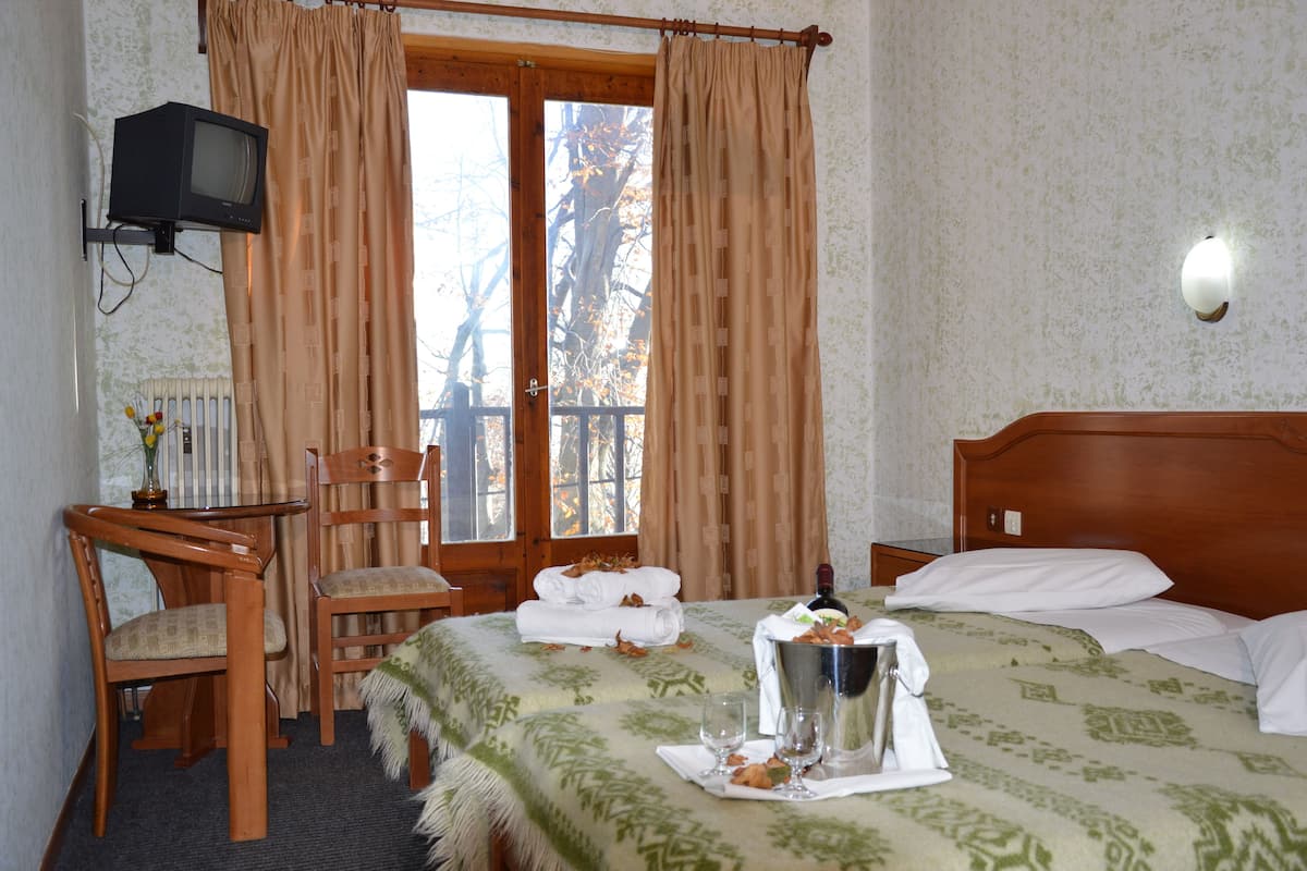 Una habitación de hotel con una cama, una silla de madera, una mesita, televisión y ventana con cortinas.