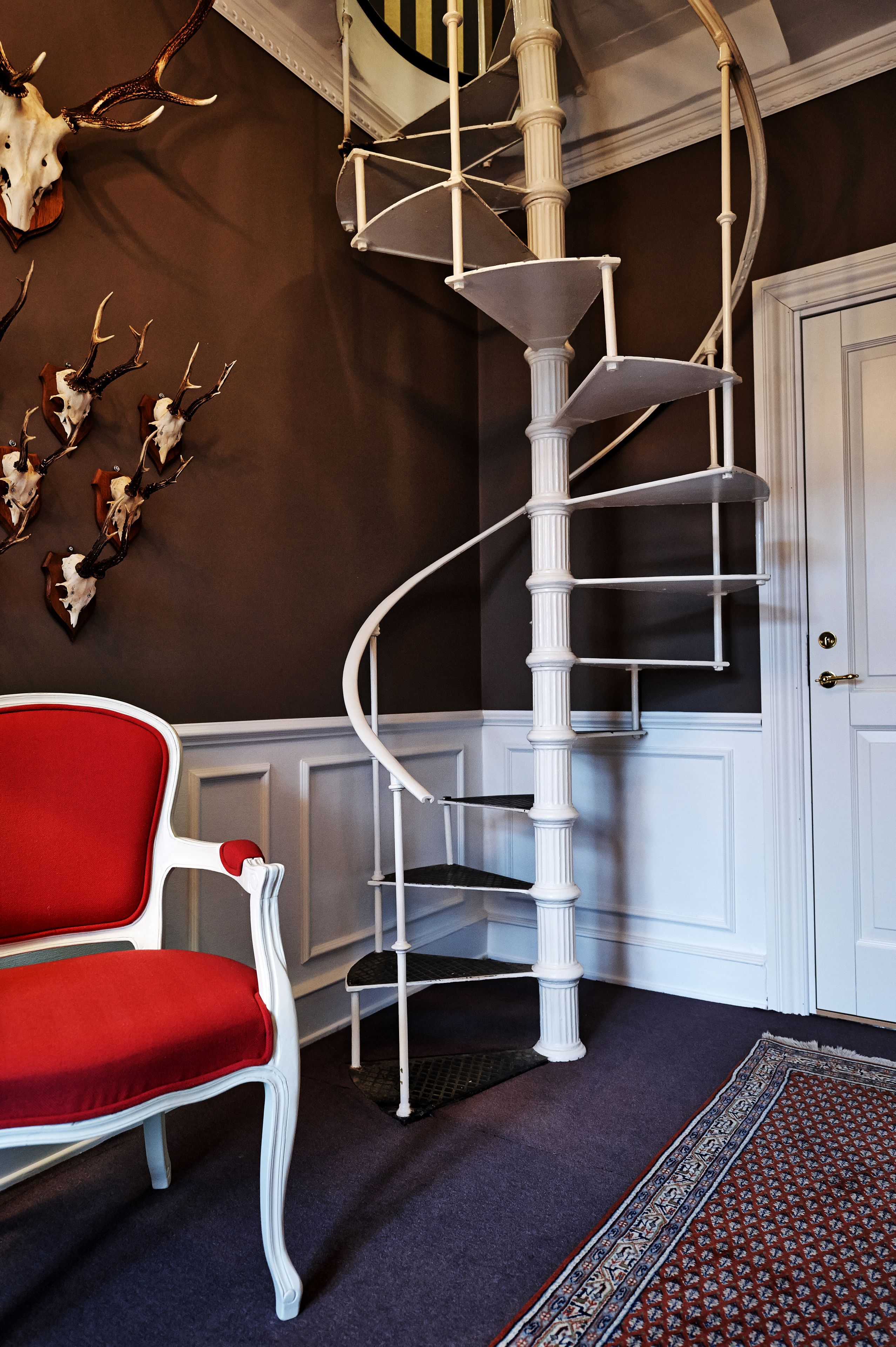 Een spiraaltrap met witte leuningen, een rode stoel en decoratieve gewei aan de muur.