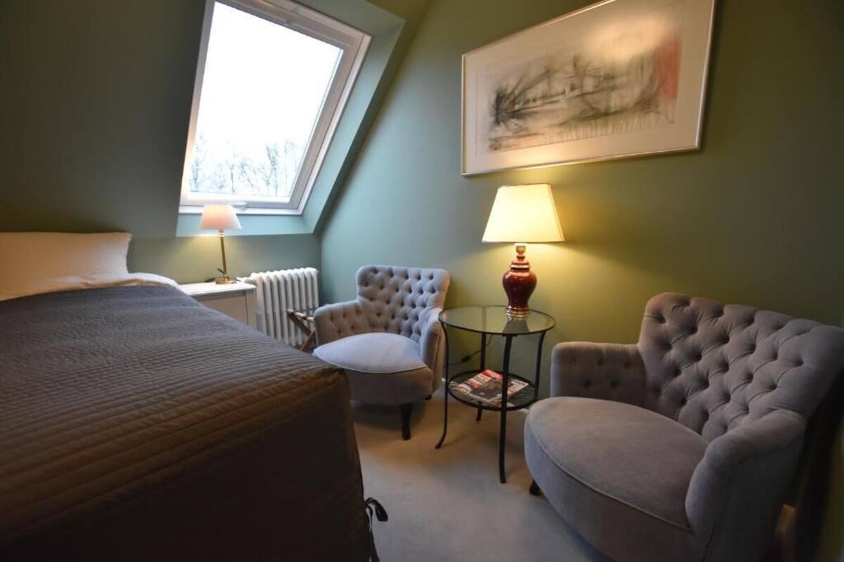 Une chambre d’hôtel avec un lit, deux fauteuils, une petite table avec une lampe, un radiateur et une fenêtre donnant sur un paysage agréable.