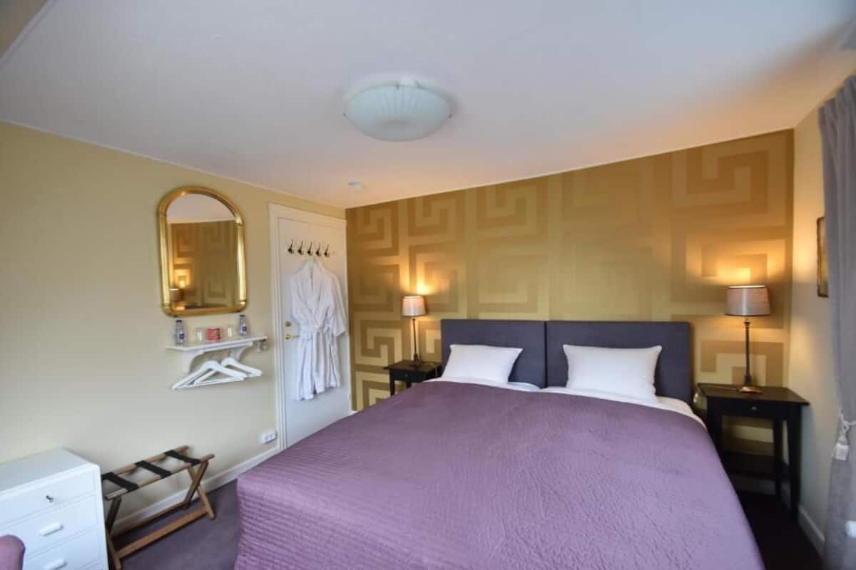 Une chambre d’hôtel avec un grand lit, deux tables de chevet avec des lampes, une armoire, un miroir et un porte-manteau.