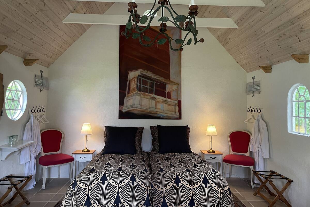 Une chambre avec un plafond haut, un lit recouvert d’une couvre-lit à motifs, deux chaises rouges, deux tables de chevet avec des lampes et un tableau représentant une maison accroché au mur.