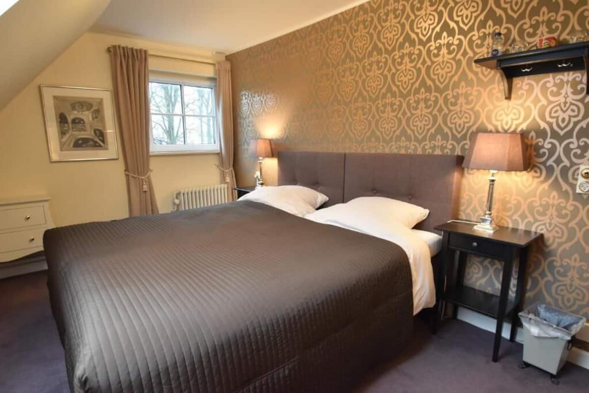 Une chambre avec un grand lit, deux lampes de chevet, un radiateur, une fenêtre avec des rideaux, un tableau encadré au mur et un papier peint à motifs.