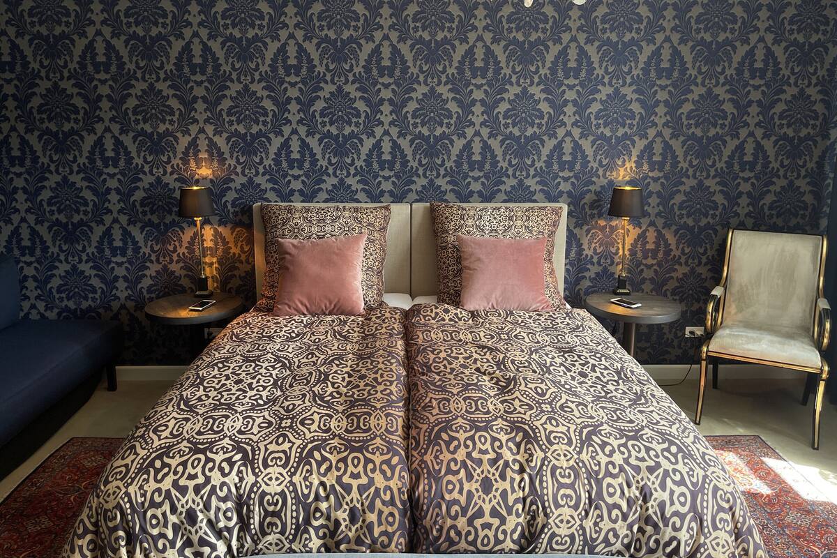 Une chambre à coucher avec une couvre-lit à motifs, deux lampes de chevet, un lustre et un fauteuil.