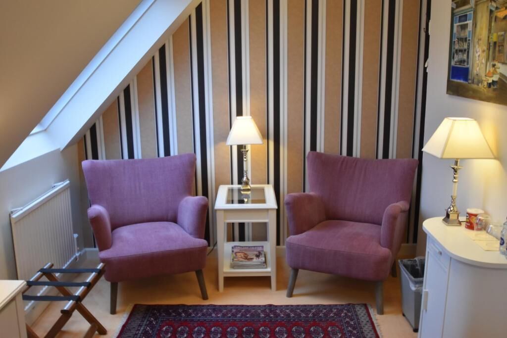 Une pièce avec deux fauteuils violets, une petite table blanche avec une lampe, un radiateur et un tapis à motifs.
