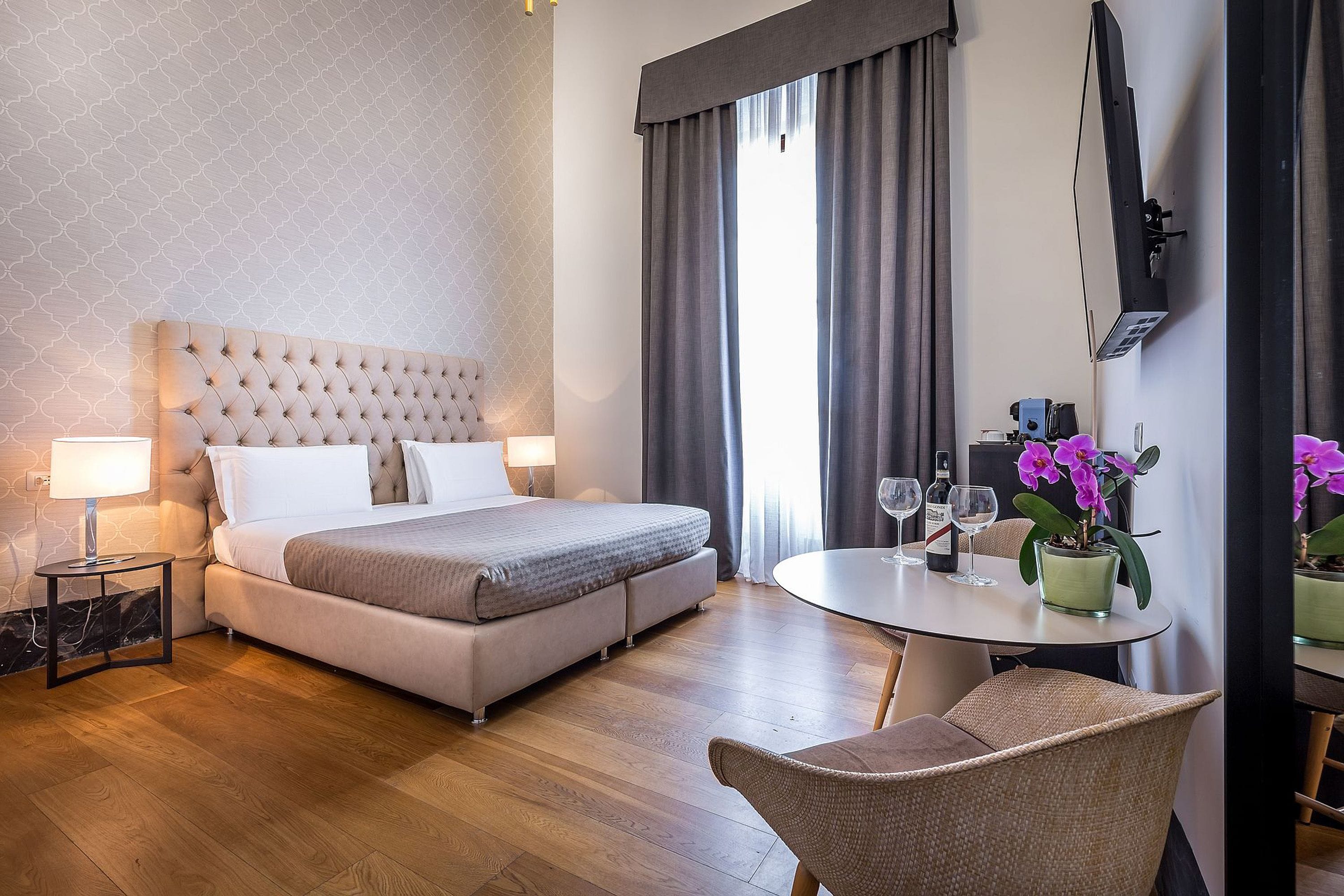 Hotellihuone, jossa on suuri sänky, yöpöytä lampun kanssa, pyöreä ruokapöytä tuoleineen ja ruukkukasvi.