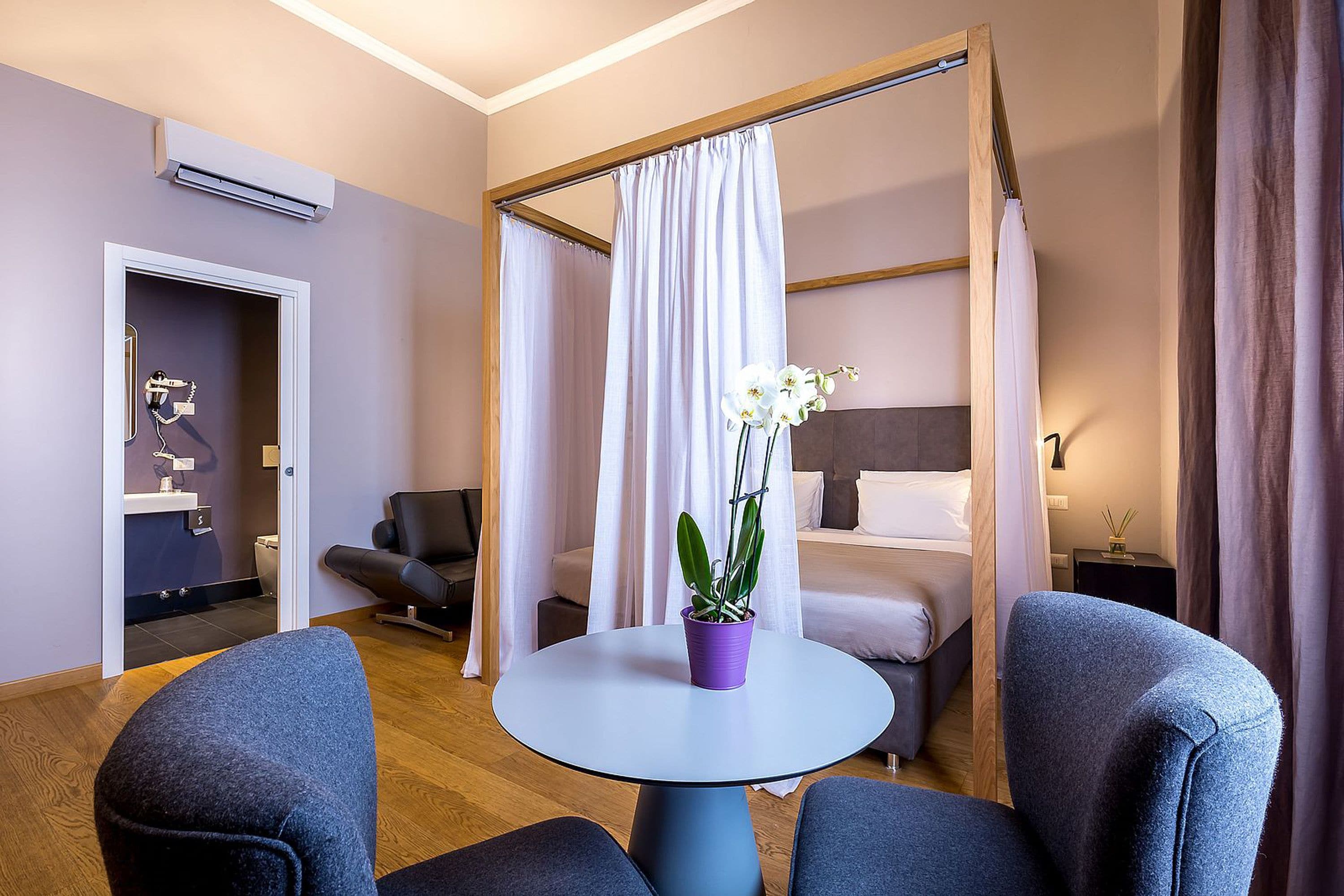 Ein Hotelzimmer mit einem Bett, einem kleinen Tisch mit Topfpflanze, einem Sessel, einem Schreibtisch und einer Dusche.