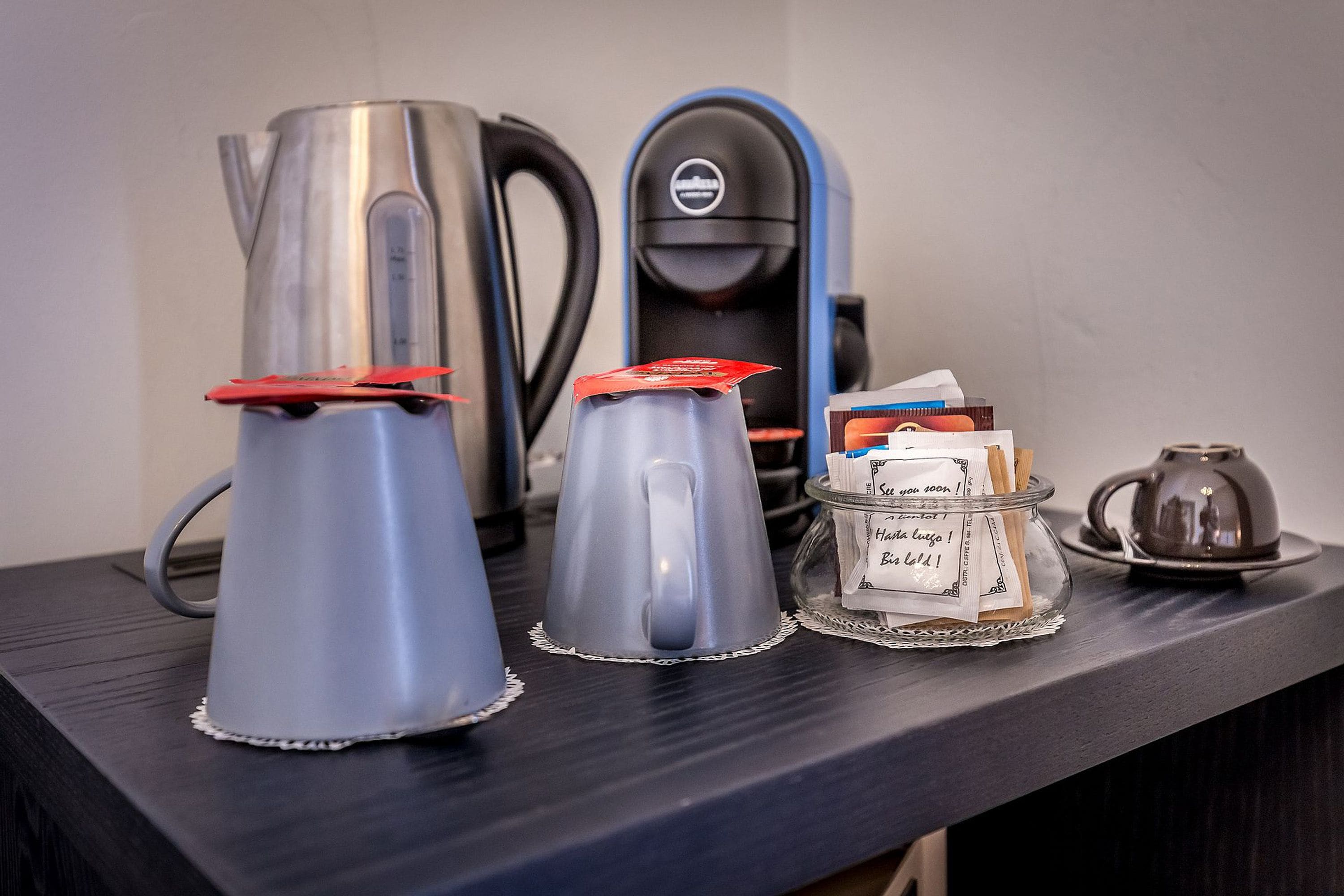 Uma estação de café com duas chaleiras, uma máquina de café e um pote de vidro com sachês de café.