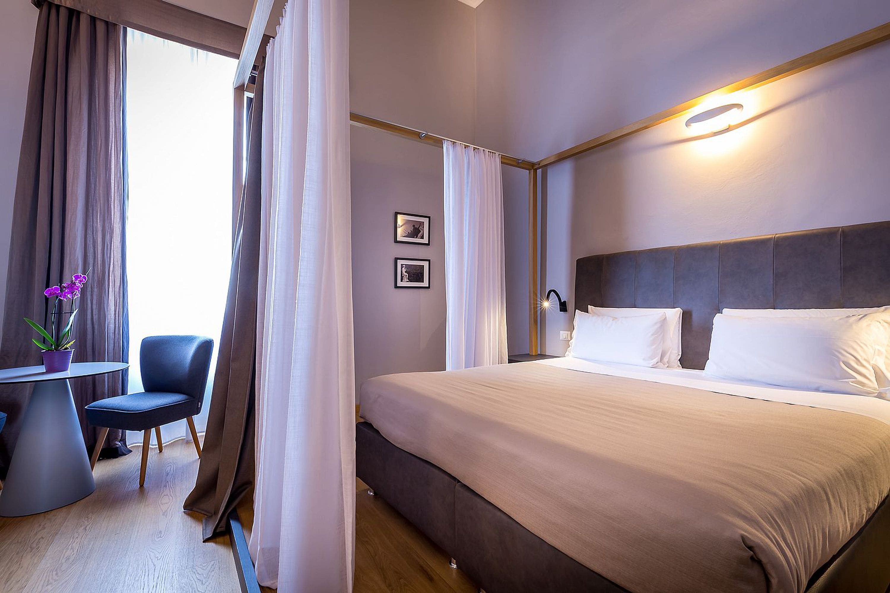 A hotel room with a large bed, a bedside table, a chair, and a small table with a potted plant.