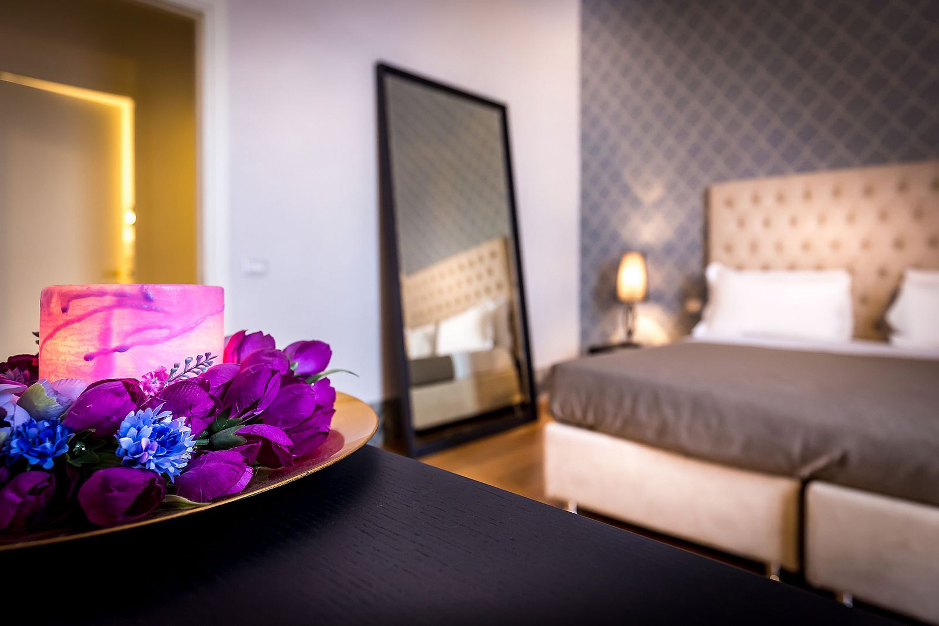 A hotel room with a bed, a bedside table with a vase of flowers, a mirror, and a wall with a patterned design.