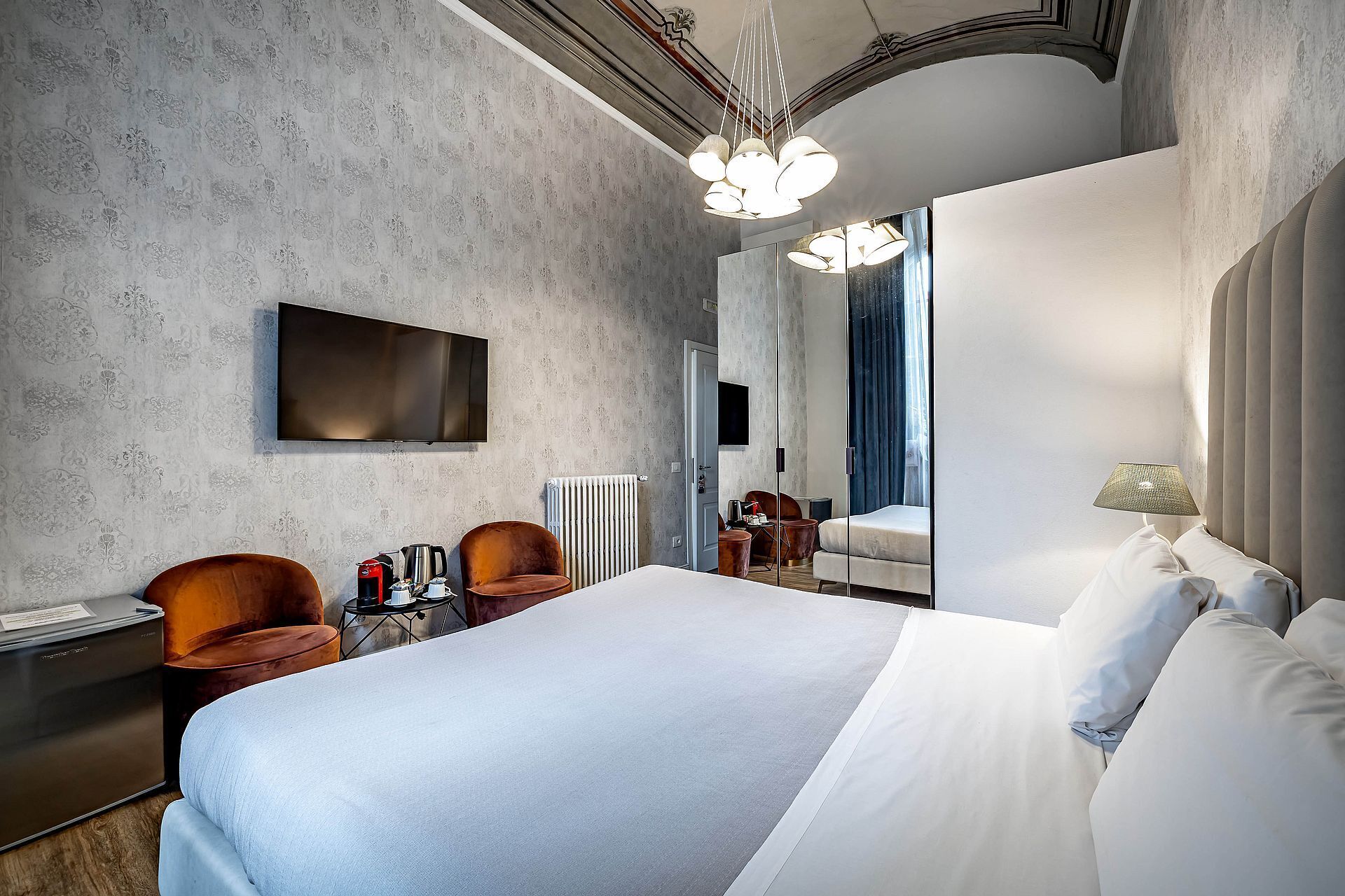 A modern hotel room with a large bed, a flat-screen TV, a coffee machine, and a seating area with chairs.