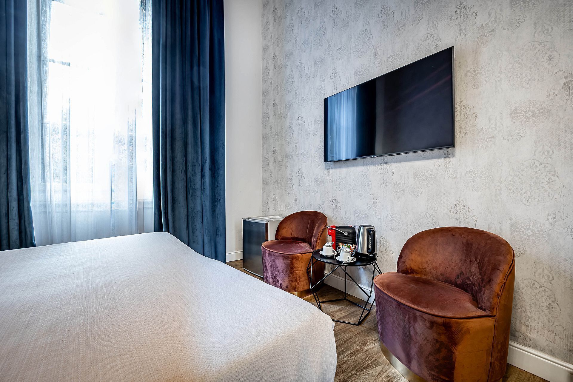 Une chambre d’hôtel avec un grand lit, deux fauteuils, une petite table avec une machine à café et un téléviseur fixé au mur.