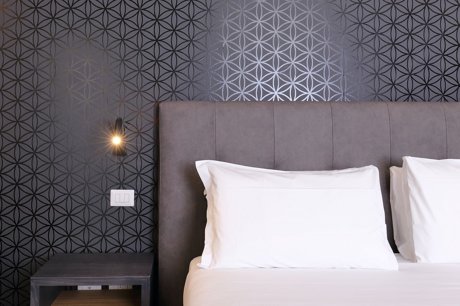 Un dormitorio moderno con papel tapiz de diseño geométrico, cabecera capitonada y mesita de noche con lámpara.