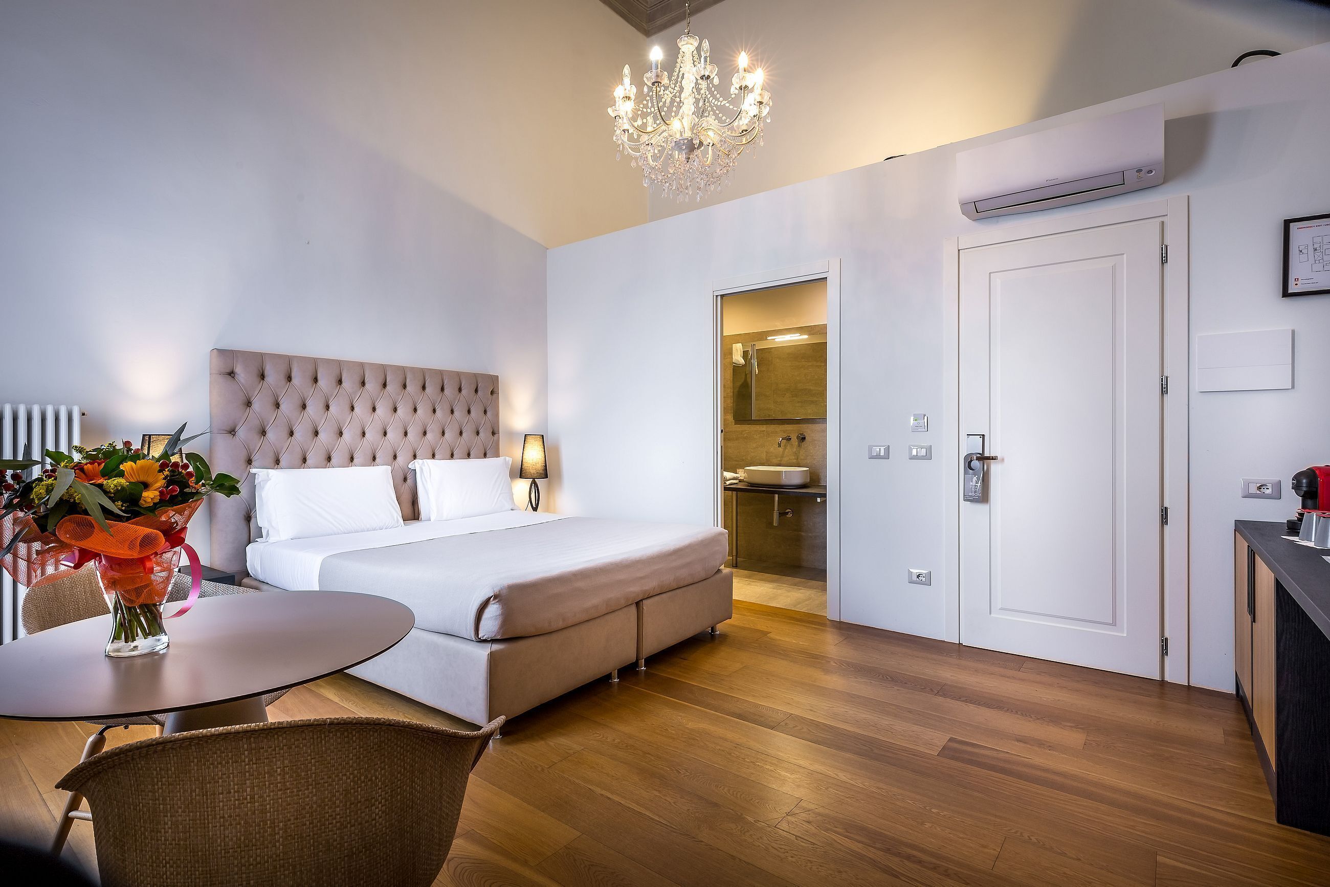 Una habitación de hotel moderna con una cama de cabecera capitonada, una mesa redonda de comedor, una araña y un baño visible a través de una puerta abierta.