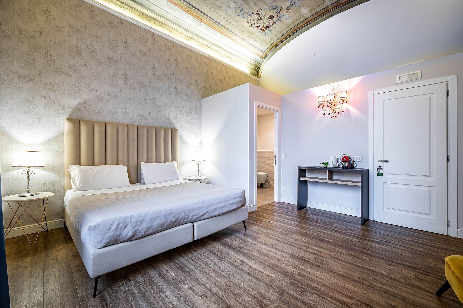 A modern hotel room with a large bed, a bedside table, a desk with a chair, and a bathroom visible through an open door.