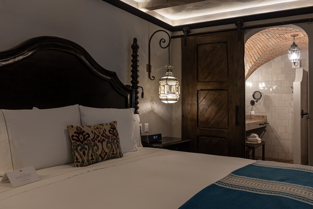 Un dormitorio con cabecera de madera, cama con almohadas estampadas, mesita de noche con un reloj y un farol colgante.