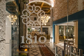 A hotel lobby with a large sign reading "Casa 1810 Hotel Boutique," featuring a checkered floor, chandeliers, and a bar area.