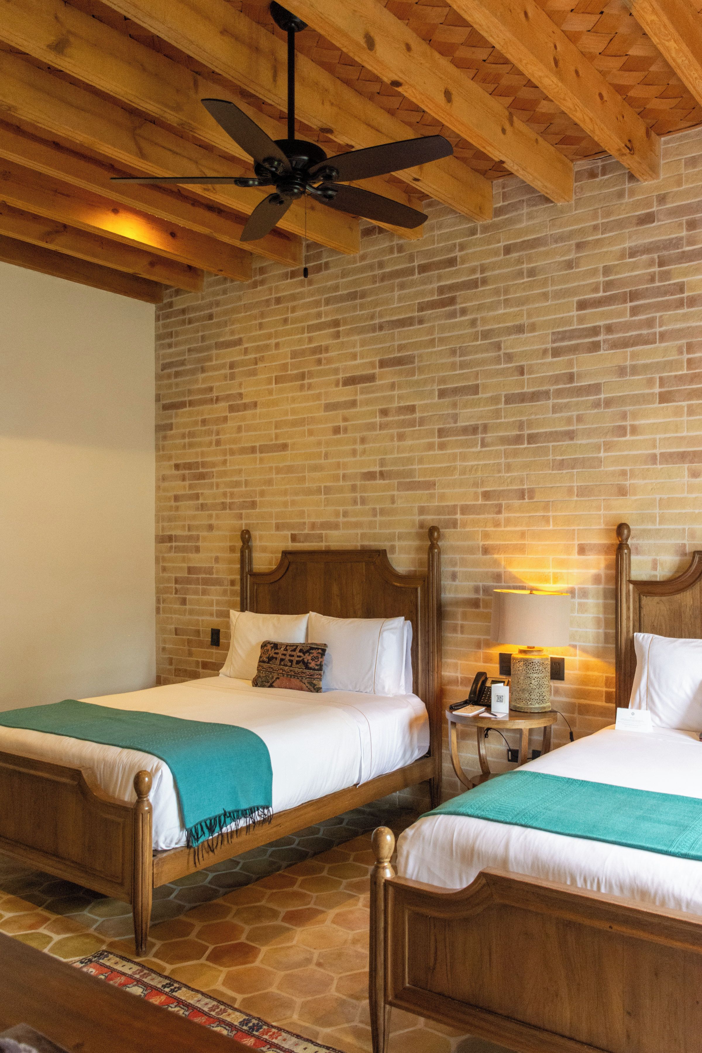 A hotel room with two wooden beds, a brick wall, a ceiling fan, and a small bedside table with a lamp.