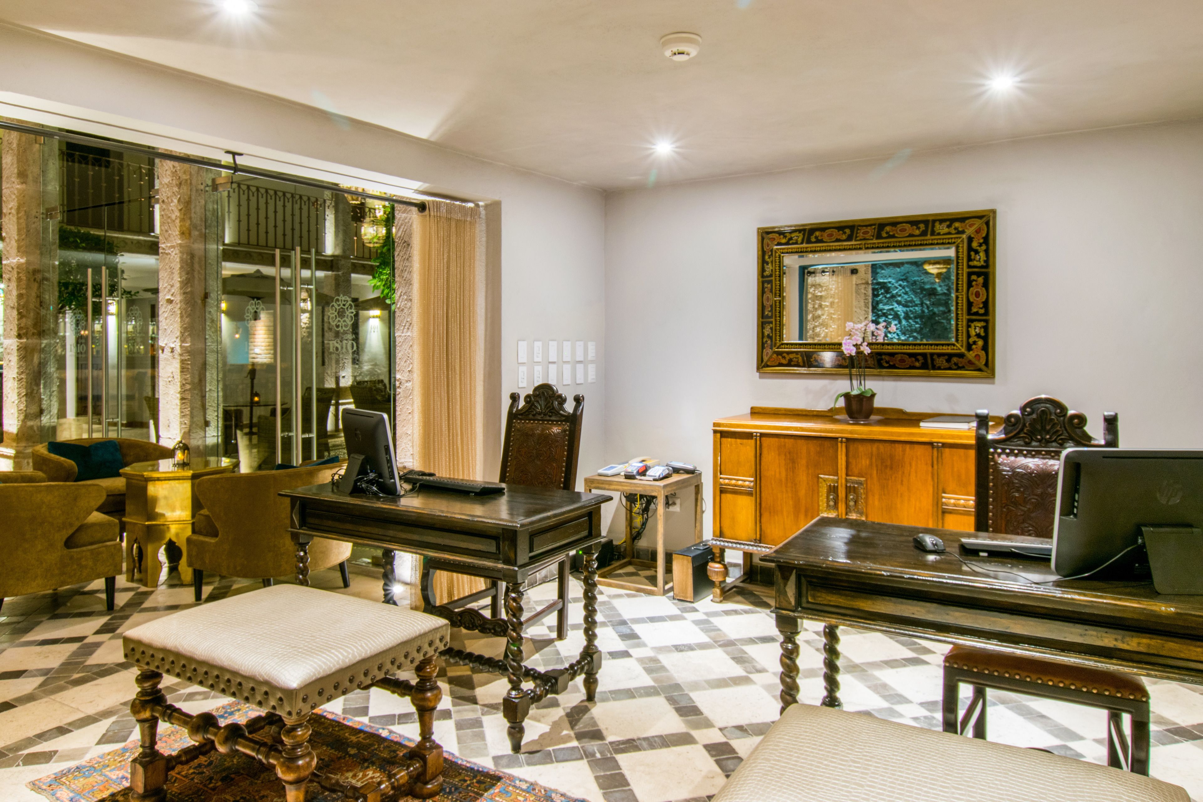 A hotel room with a checkered floor, a desk with a computer, a chair, a sofa, and a large mirror on the wall.