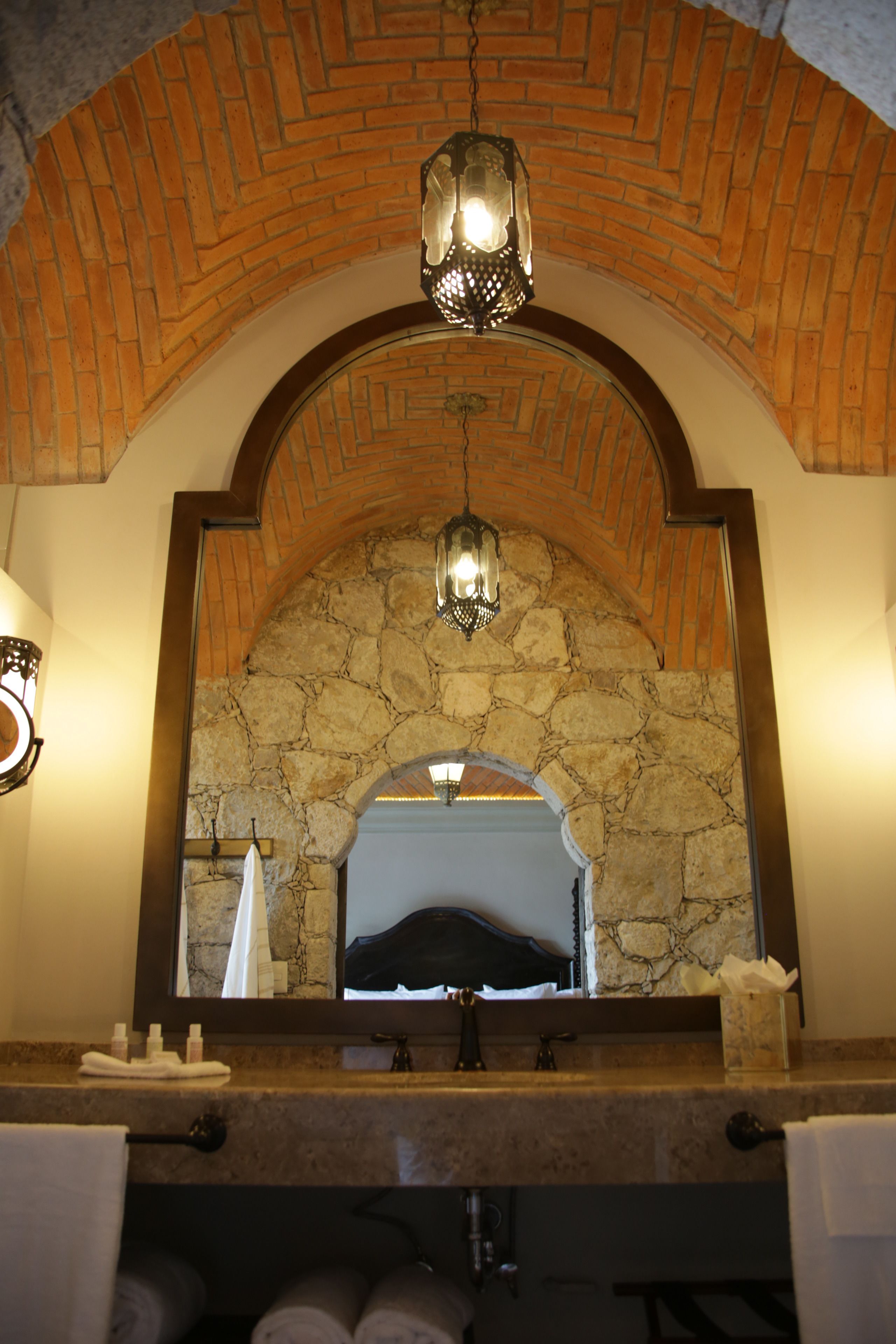 Casa de banho com arco de pedra, candeeiro pendente e espelho a refletir uma cama.