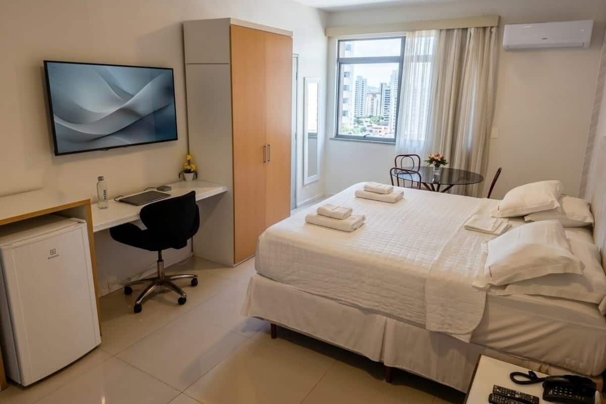 Ett hotellrum med en säng, ett skrivbord med en stol, en TV, ett litet kylskåp, ett fönster med gardiner och en luftkonditioneringsanläggning.