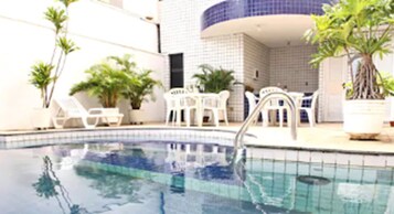 Piscina com deck revestido, rodeada por vasos de plantas e mobiliário de exterior branco.