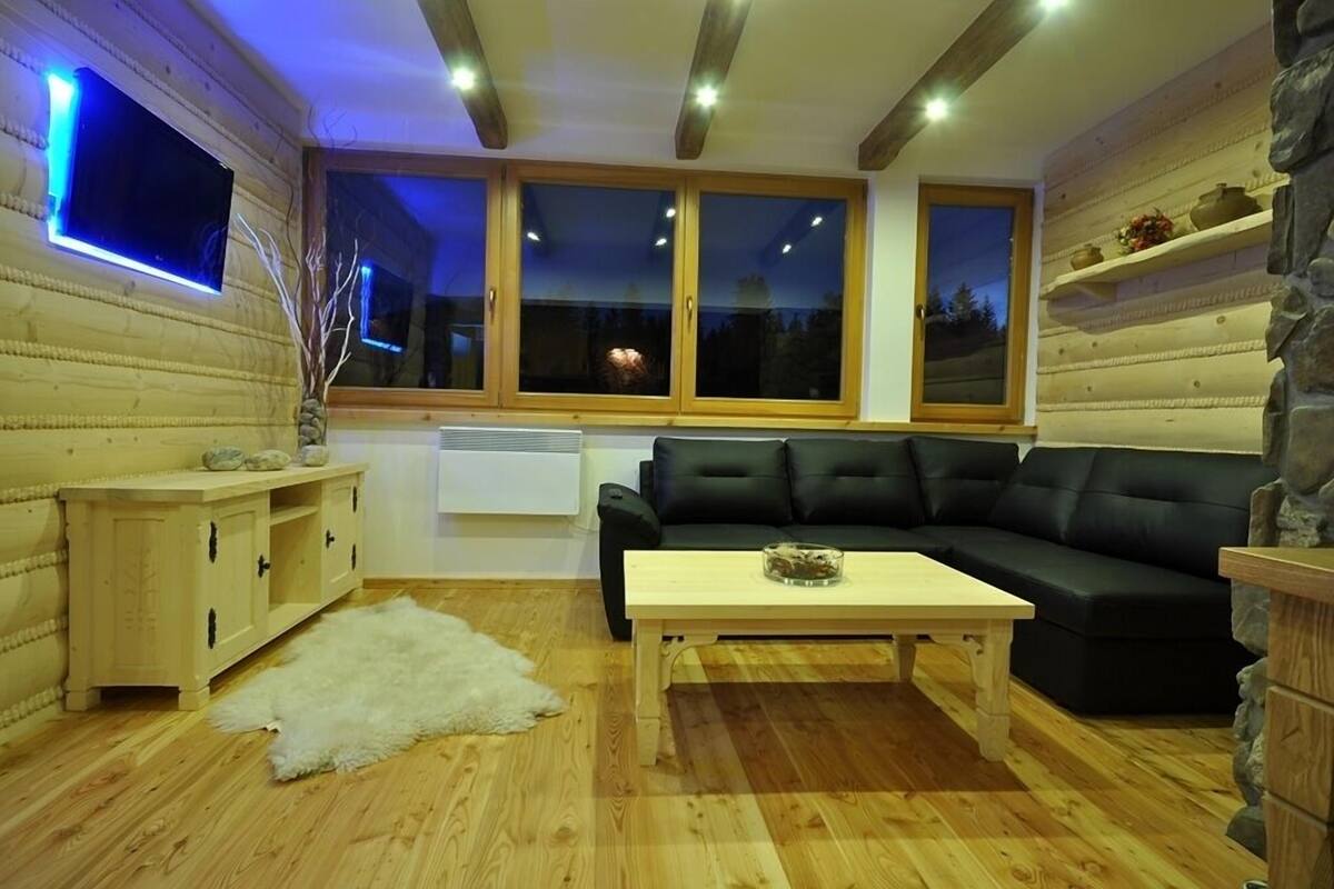 Ein gemütliches Wohnzimmer mit Holzboden, einer schwarzen Ledersofa, einem Holztisch und einem weißen Teppich. An einer Seite befinden sich Holzschränke und eine Steinwand.