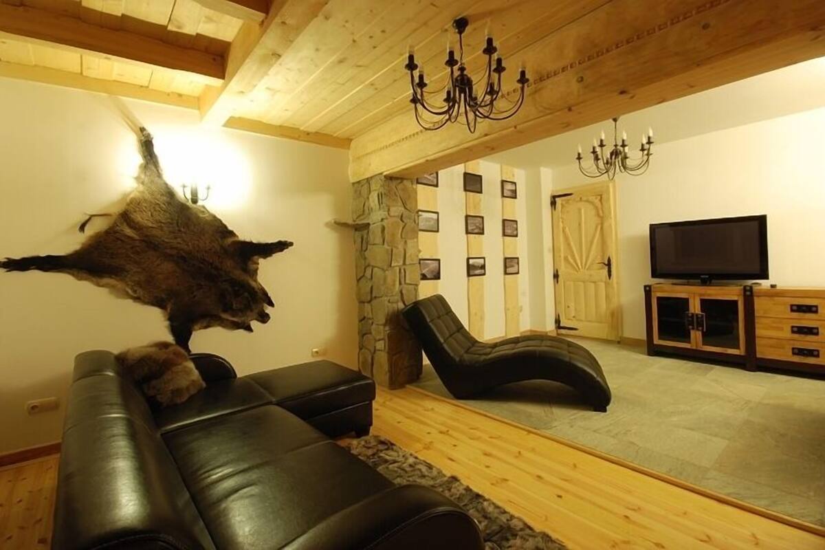 Ein Wohnzimmer mit einer Steinwand, einem schwarzen Ledersofa, einem Liegestuhl, einem Fernseher und einer Chandelüster.