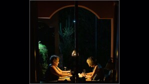 Dos personas sentadas a una mesa dentro de una habitación, con una ventana, una lámpara y una planta visibles desde el exterior.