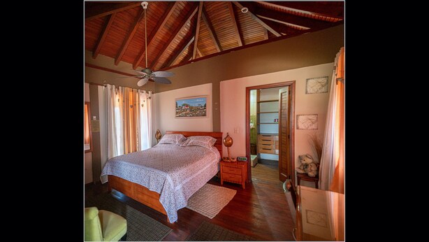 Un dormitorio con techo de madera, una cama con colcha estampada, una mesita de noche, una silla y una puerta que da a otra habitación.