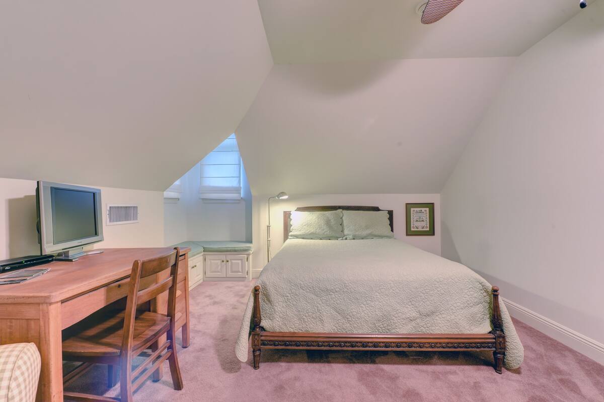 A bedroom with a bed, a desk, a chair, a television, and a window.