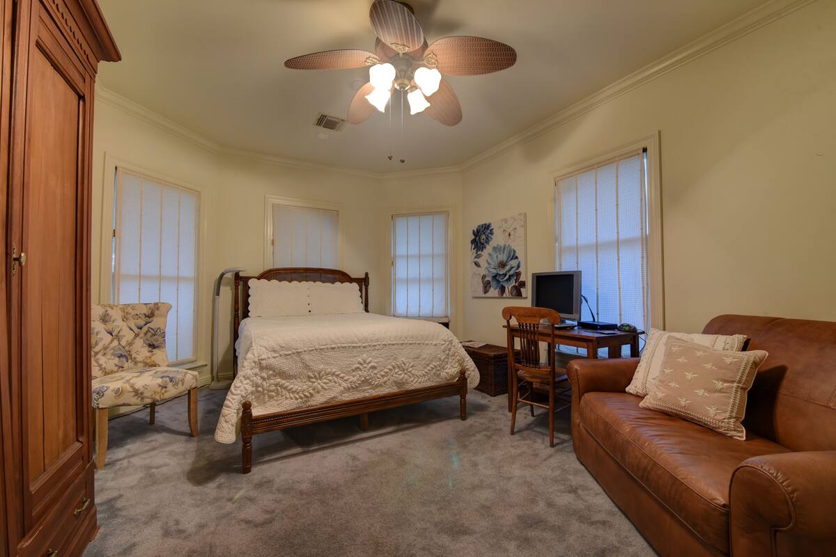 A bedroom with a bed, a wooden wardrobe, a brown leather sofa, a wooden chair, and a ceiling fan.