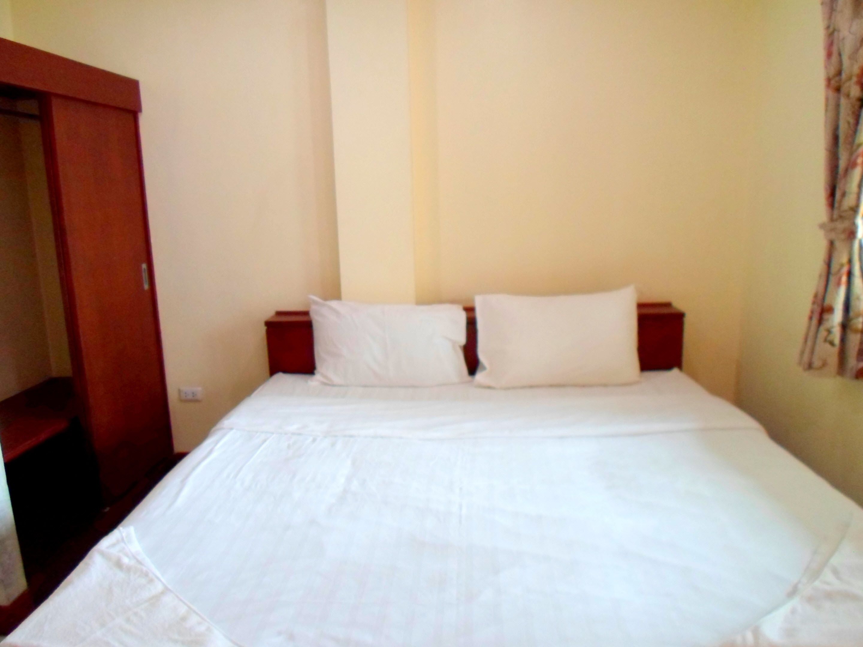 A standard hotel room with a single bed, two pillows, a wooden nightstand, and a wardrobe.