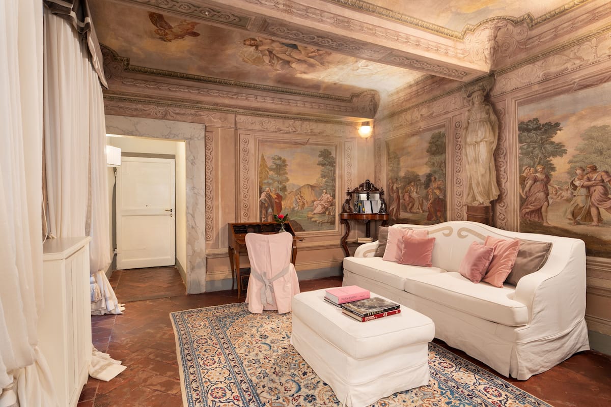 Una habitación con un sofá blanco, un reposapiés a juego, una alfombra estampada y un techo con frescos clásicos.