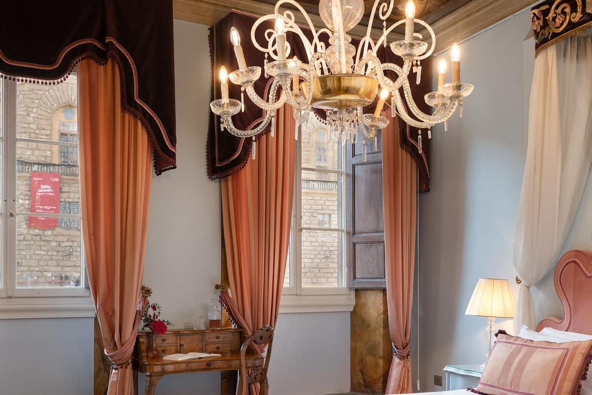 Un dormitorio con una cama grande, un candelabro, un escritorio con una silla y una ventana con cortinas.