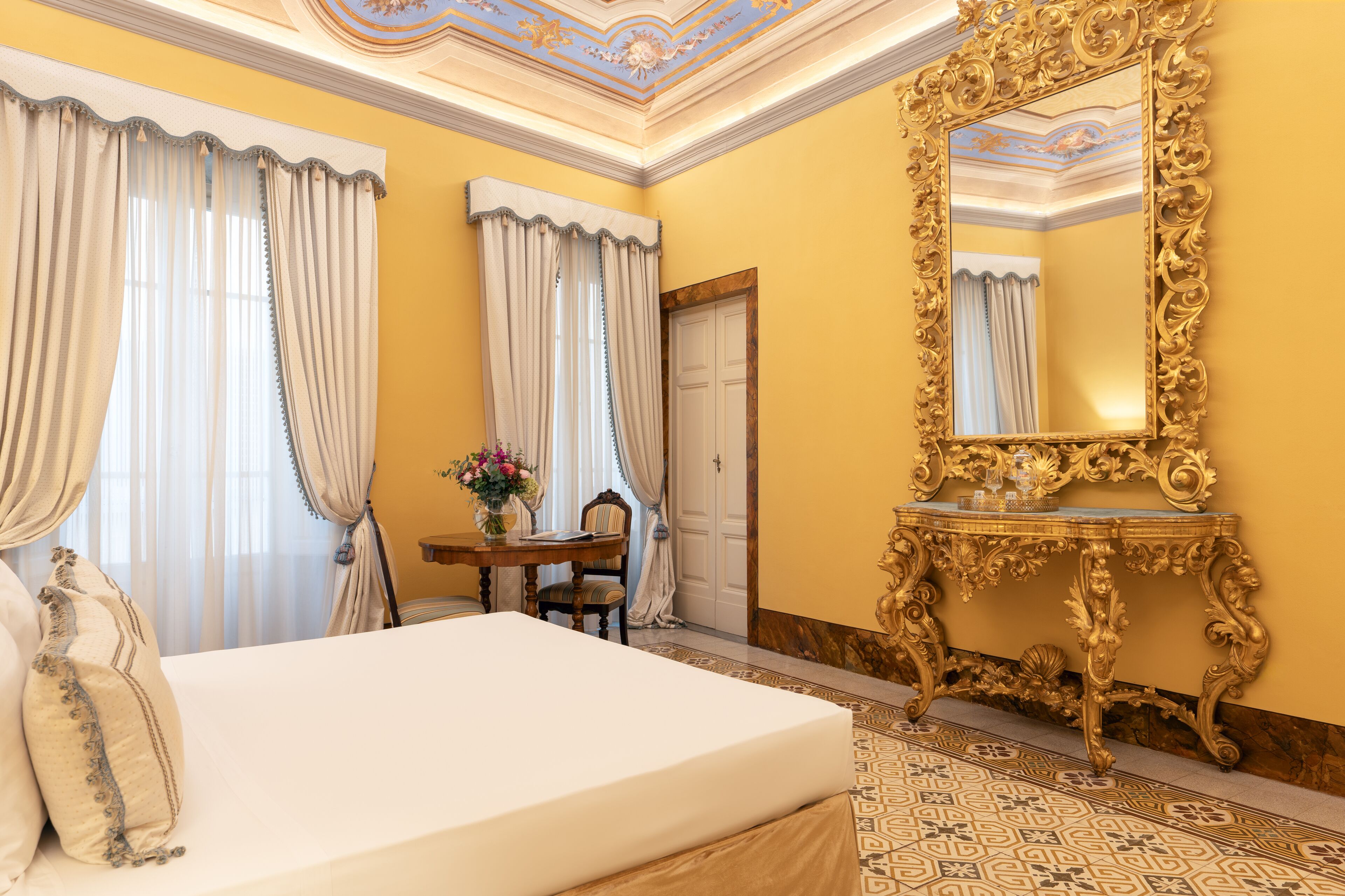 Habitación de hotel con una cama grande, una mesa con un florero, una silla y un espejo con marco dorado.
