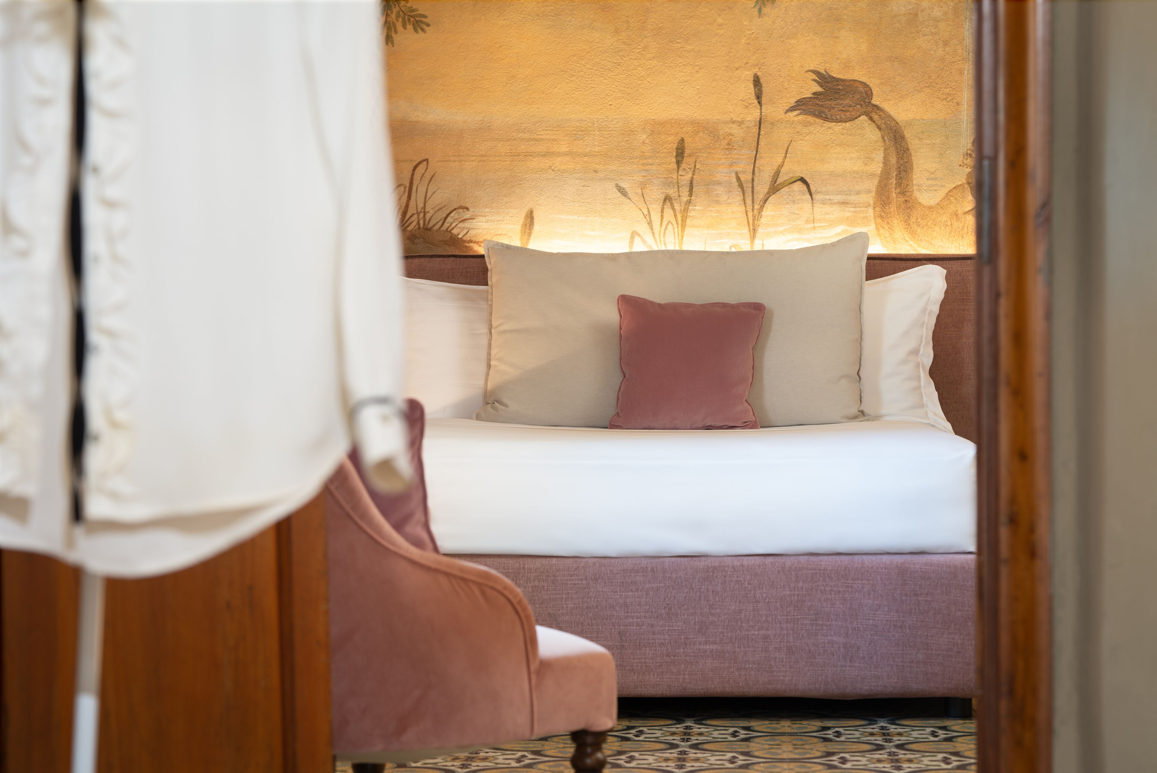 A neatly made bed with white and pink pillows, a pink chair, and a wall with a nature-themed painting.