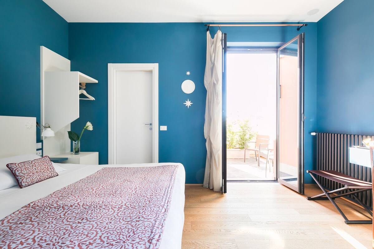A bedroom with a bed, a white nightstand, a white dresser, a white wall with a circular decoration, and a door leading to a balcony.