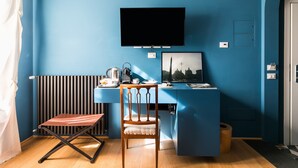 Ein kompaktes Zimmer mit blauer Wand, einem flachbildfernseher, der über einem Schreibtisch angebracht ist, einem Holzstuhl, einem Heizkörper und einem kleinen Holzschemel.