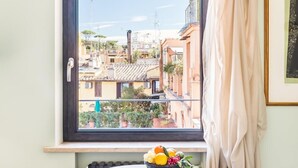 Ein Zimmer mit Fensterblick auf eine Stadtlandschaft, einem kleinen runden Tisch mit Obstschale und einem Heizkörper darunter.