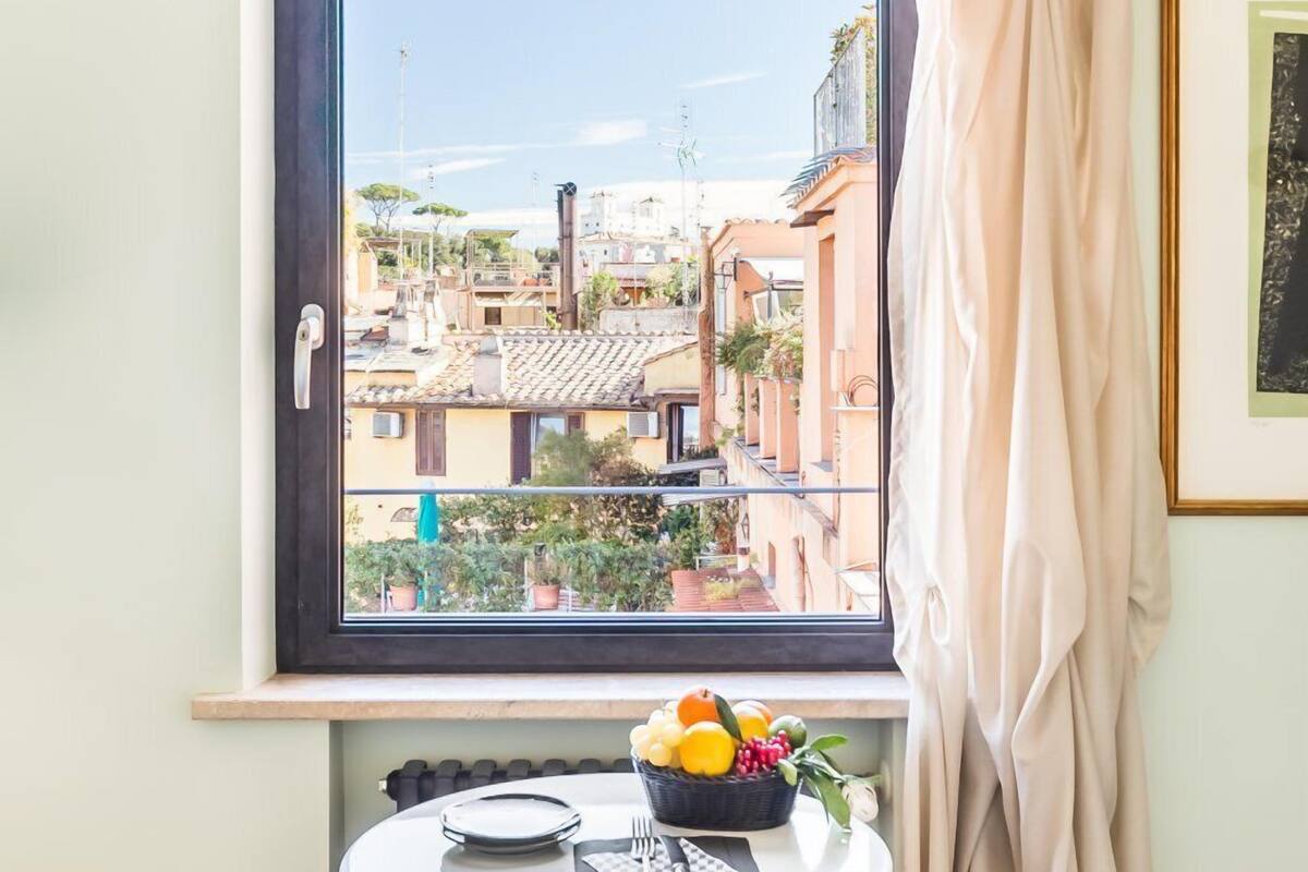 A room with a window overlooking a cityscape, a small round table with a fruit bowl, and a radiator below.