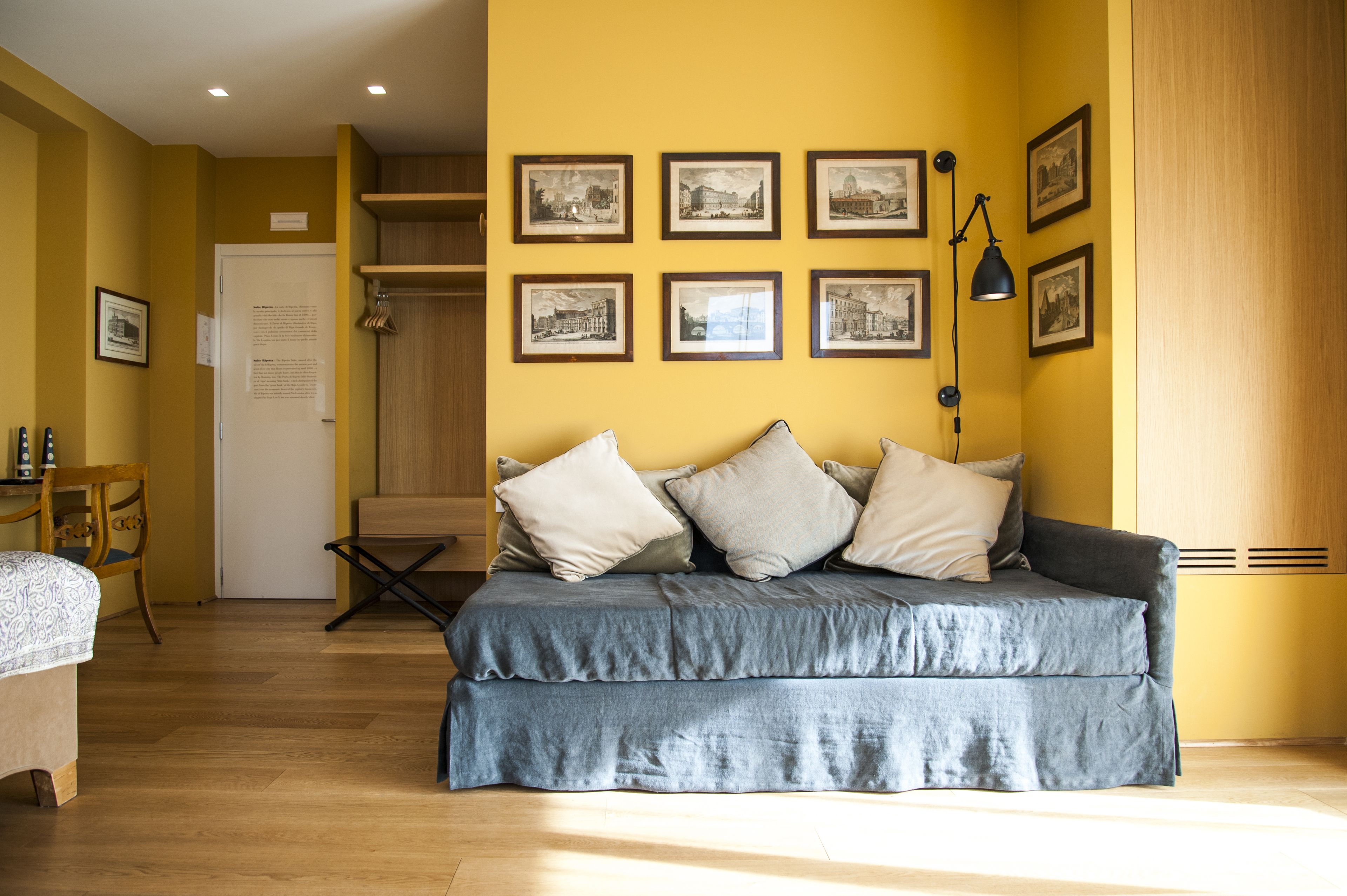 Une pièce comprenant un canapé, une chaise en bois, une petite table et un lit. Des tableaux encadrés ornent le mur, et il y a une porte avec une pancarte.