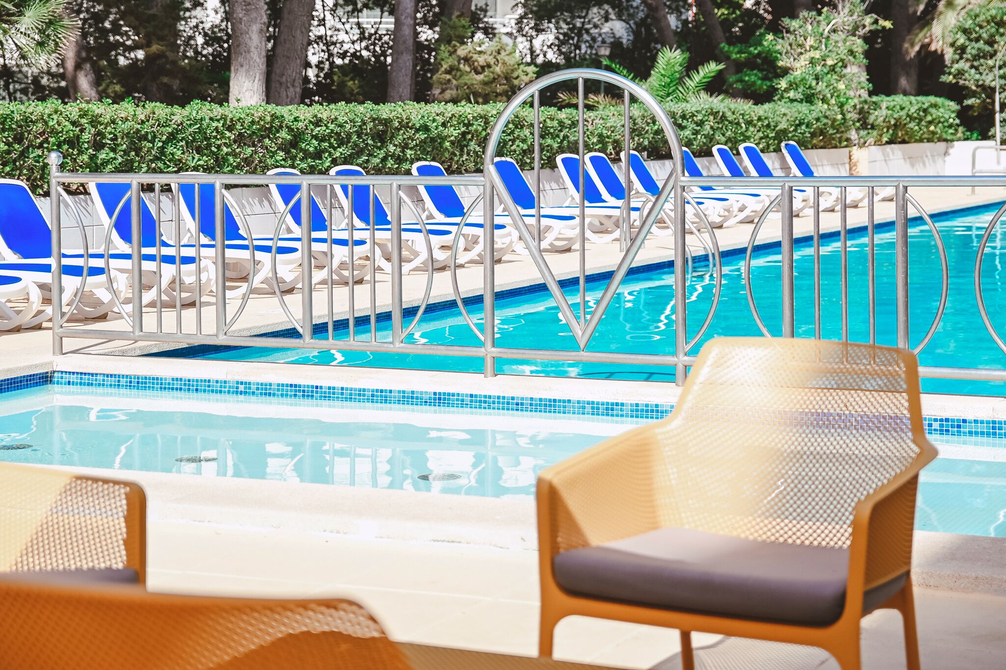 Un área de piscina con sillones de descanso azul y blanco, una escalera metálica para la piscina y una silla de mimbre al frente.