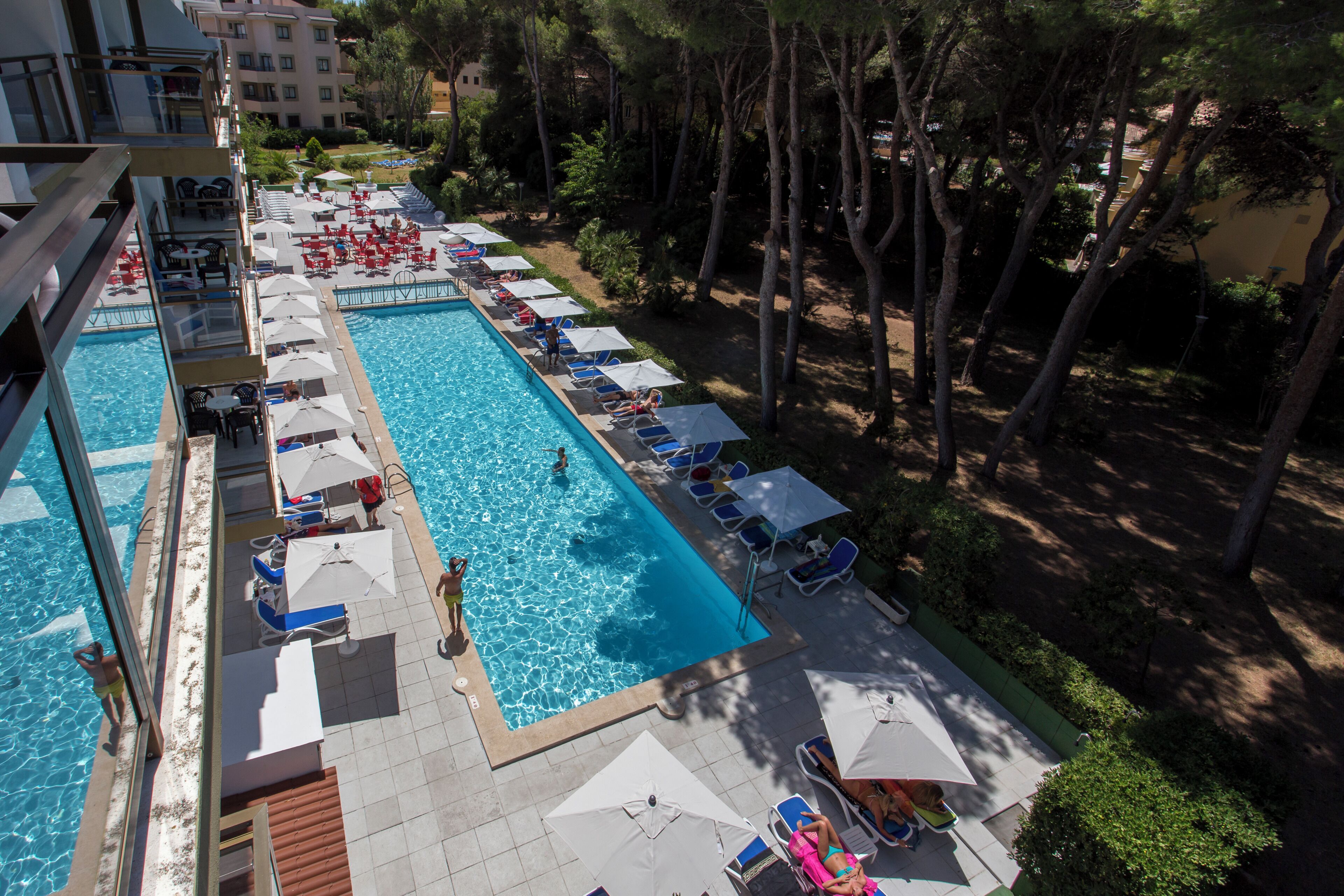 A swimming pool with sun loungers and umbrellas, surrounded by trees and a building in the background.