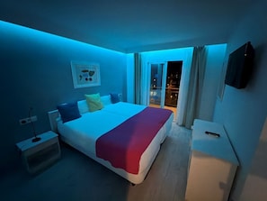 A hotel room with a bed, a nightstand, a TV, and a view of the outside.