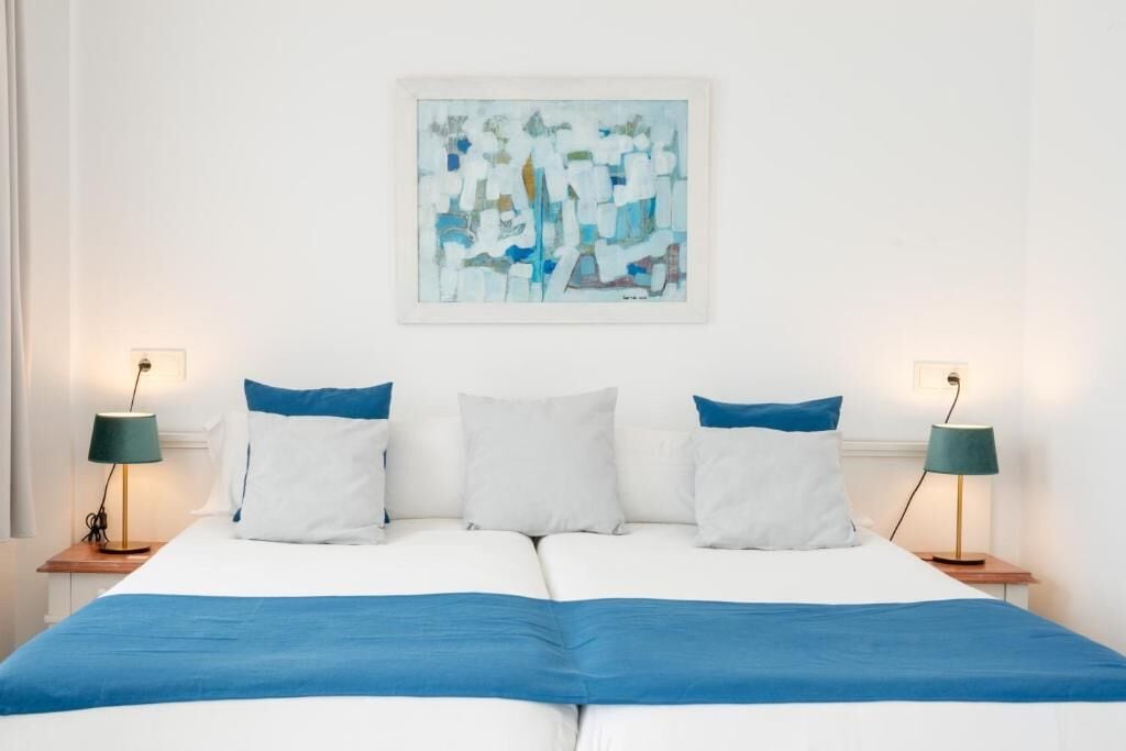 Cama doble con una colcha azul, almohadas blancas y dos mesitas de noche con lámparas.