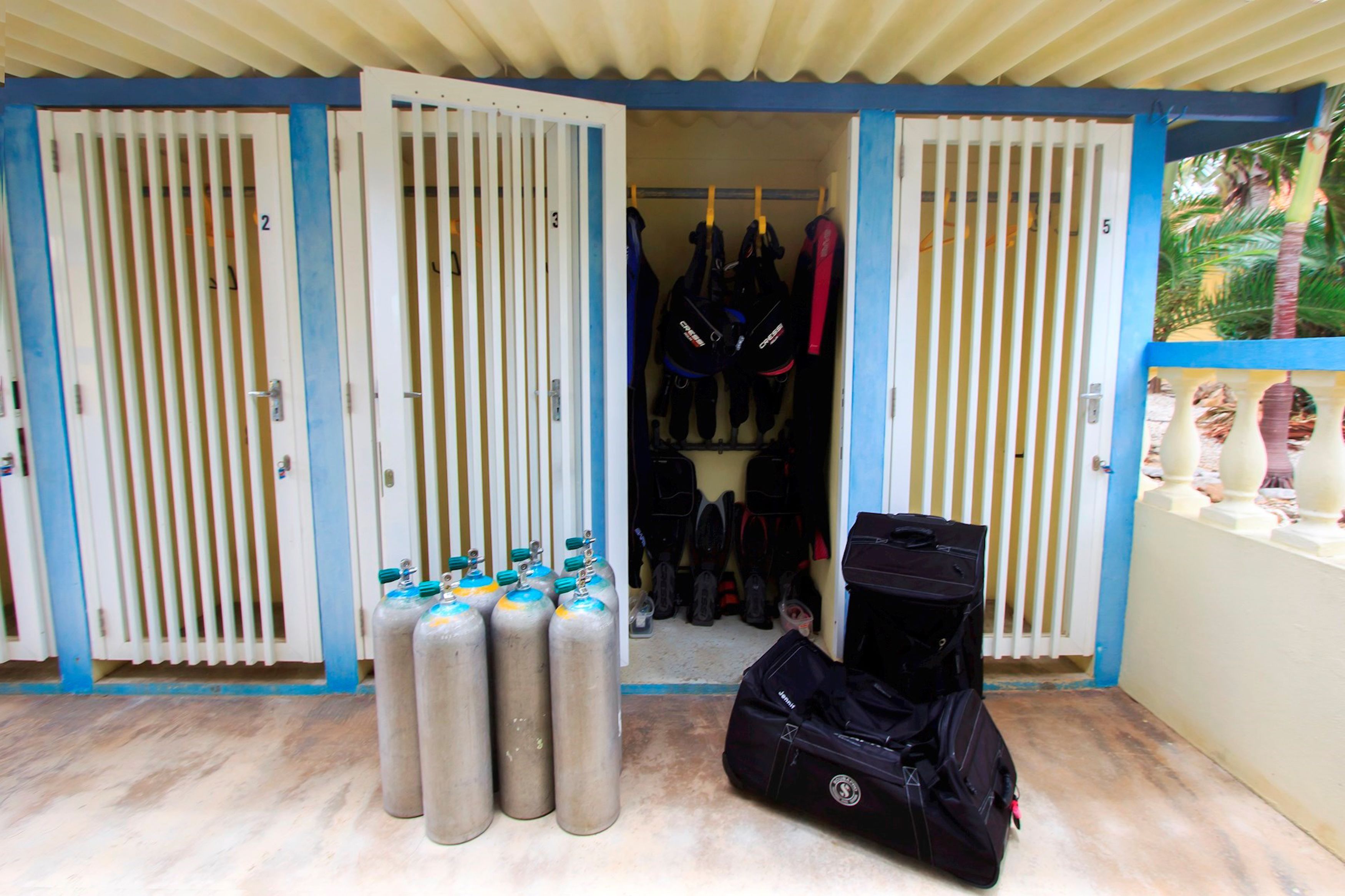 A room with a blue and white color scheme, featuring a door with numbered panels, a rack of scuba gear, and several scuba tanks.