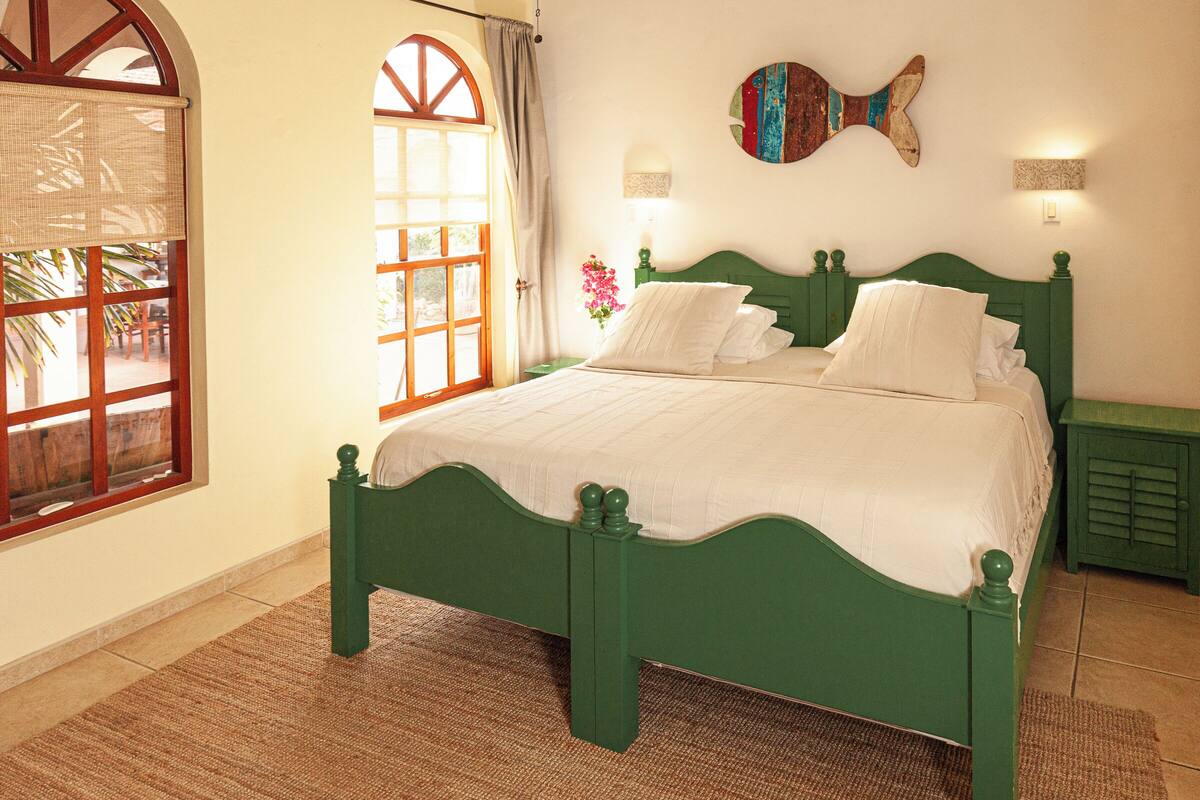 A green bed with white linens in a room with a large window and a fish-shaped wall decoration.
