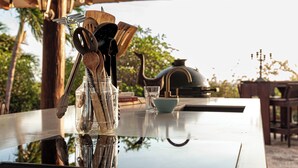 A table with kitchen utensils and a kettle, reflecting on a glass surface, under a wooden structure with hanging lights. - Goood Resort (Kralendijk)